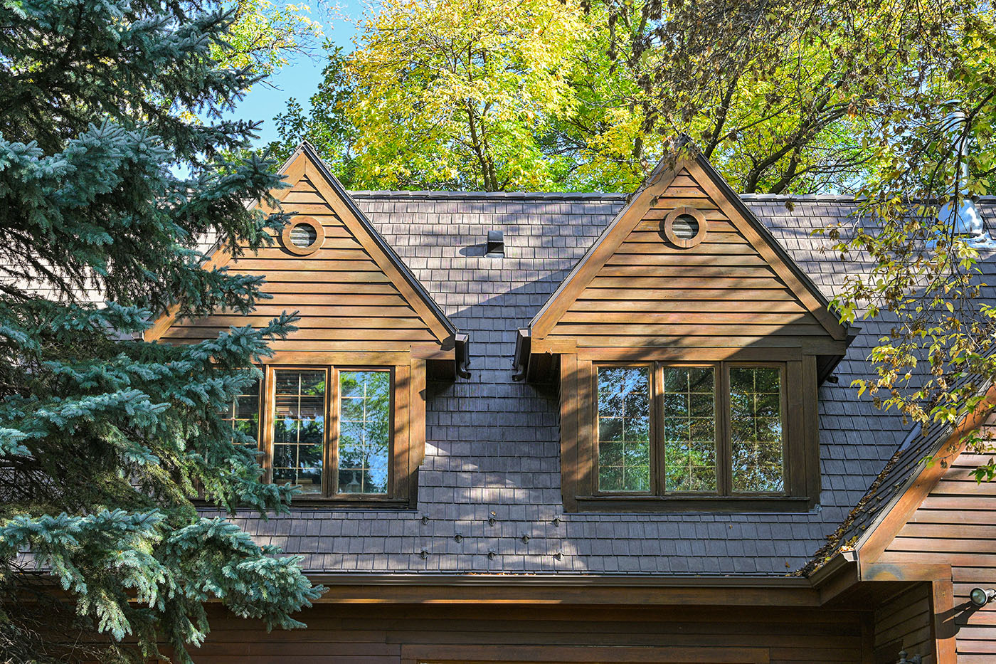 Twin dormers with round windows set into a grey slate-look shingle roof among trees.