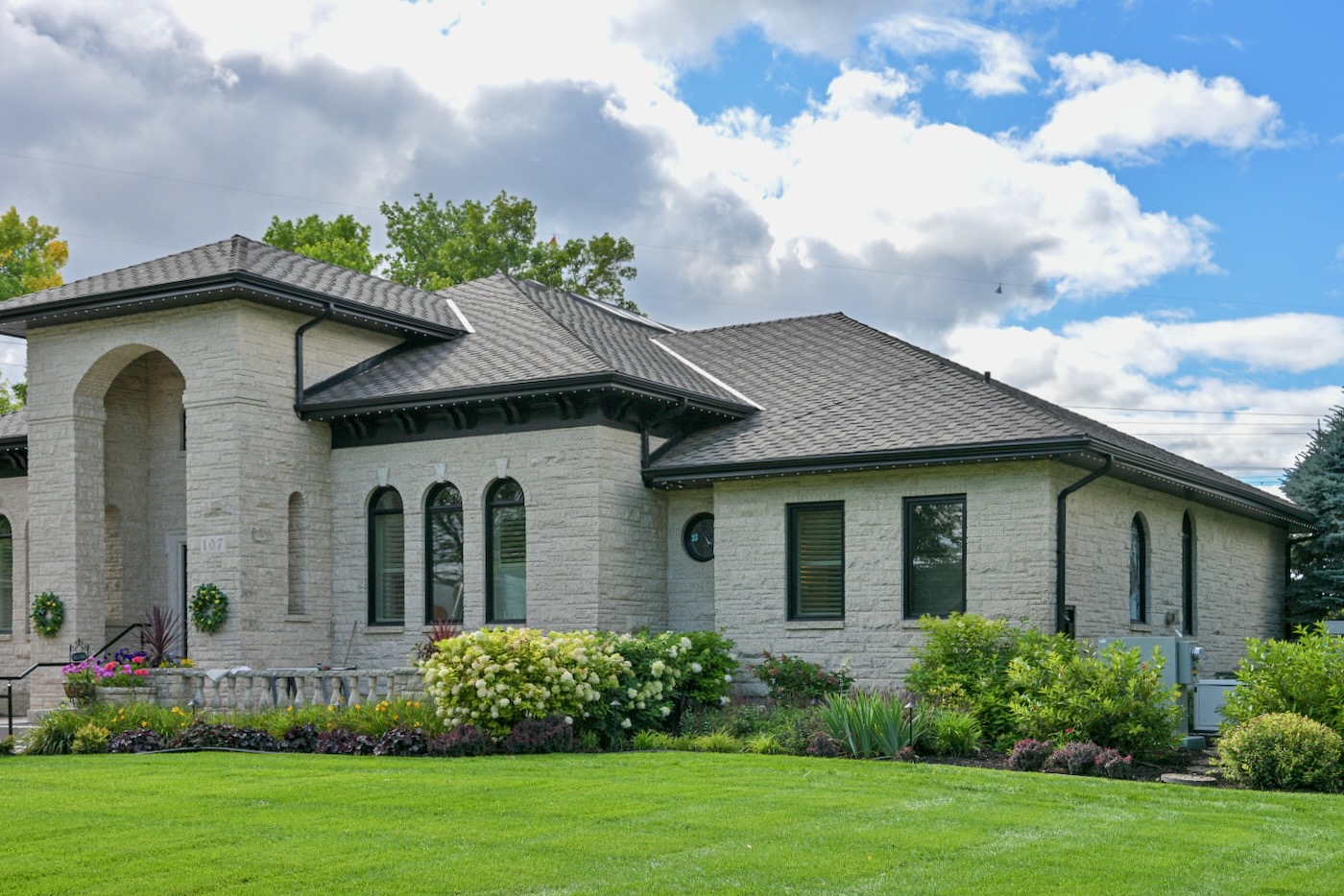 Stone home with dark shingle roof and colourful landscaped front lawn